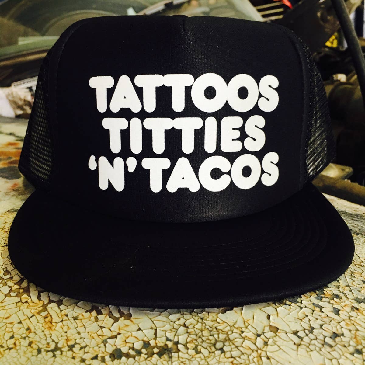 Black cap with white text 'TATTOOS TITTIES 'N' TACOS' on a textured surface