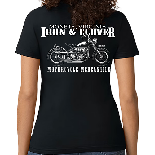 A woman wearing a black crew neck t-shirt with a graphic of a motorcycle and the text 'IRON & CLOVER MONETA, VIRGINIA MOTORCYCLE MERCANTILE'. The shirt is made by Gildan.