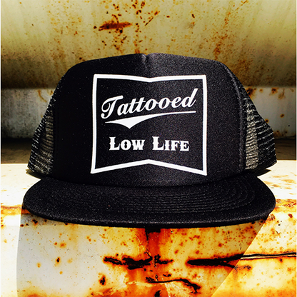 Black trucker hat with 'Tattooed Low Life' text on a rusty metal surface