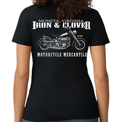 A woman wearing a black crew neck t-shirt with a graphic of a motorcycle and the text 'IRON & CLOVER MONETA, VIRGINIA MOTORCYCLE MERCANTILE'. The shirt is made by Gildan.